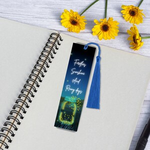 May include: A blue bookmark with a white tassel. The bookmark features a dark blue background with a white jar filled with glowing fireflies. The text on the bookmark reads "Fireflies, Sunshine and Rainy days".