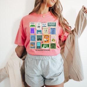 Peut inclure: T-shirt corail avec un collage de timbres-poste de style vintage. Les timbres présentent divers motifs et textes, dont les mots "BITTERSWEET SYMPHONY" en bas. Le t-shirt a des manches courtes et une coupe décontractée.