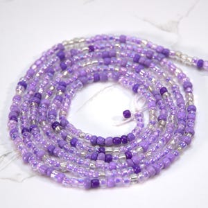 Könnte beinhalten: Eine violette und silberne Perlen-Taillenkette mit schillernden Perlen. Die Kette ist auf einer weißen Oberfläche aufgerollt.