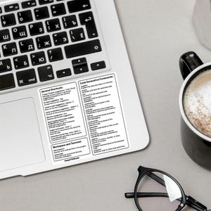 Może przedstawiać: Czarno-biała naklejka z listą og&oacute;lnych skr&oacute;t&oacute;w klawiszowych i poleceń generowania kodu dla aplikacji do programowania komputerowego. Naklejka znajduje się na klawiaturze laptopa z filiżanką kawy i okularami w pobliżu.