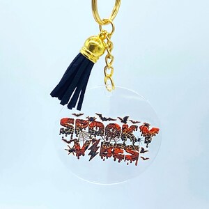 May include: A clear round keychain with a black tassel and a gold chain. The keychain has a printed design of the words "Spooky Vibes" in orange and black with bats, spiders, and spiderwebs.