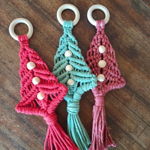 Könnte beinhalten: Drei handgefertigte Makramee-Weihnachtsbaumschmuckstücke mit Holzperlen und Quasten. Die Bäume sind rot, türkis und rosa.