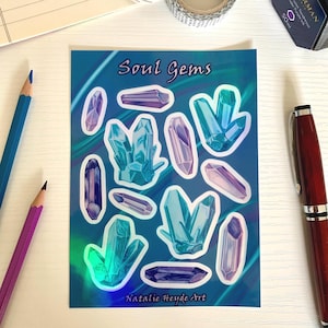 Könnte beinhalten: Eine Bogen mit holografischen Aufklebern mit 12 bunten Kristallformen. Die Aufkleber befinden sich auf einem blauen Hintergrund mit dem Text "Soul Gems" und "Natalie Heyde Art".