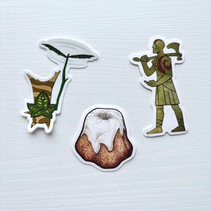 Könnte beinhalten: Eine Sammlung von drei gestanzten Aufklebern. Einer zeigt eine stilisierte Pflanze mit einem großen weißen Blatt und grünen und braunen Akzenten. Ein weiterer zeigt einen Gugelhupf mit weißer Glasur. Der dritte ist eine Kriegerfigur mit einer Axt.