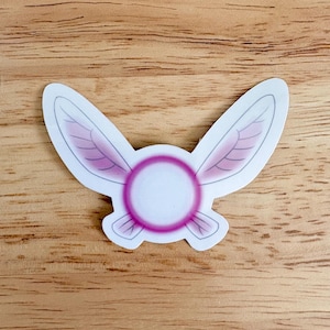 May include: A white sticker with a pink and purple fairy wing design. The sticker features a central circle with a pink outline and two stylized wings with purple shading and gray outlines. The sticker is on a wooden surface.