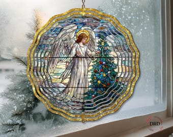 May include: A stained glass-style Christmas ornament featuring an angel with a halo, standing near a decorated Christmas tree. The ornament has a gold-colored, layered, wavy border. The angel is wearing a white robe. The background is a winter scene.