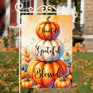 May include: A garden flag with a design of stacked pumpkins in orange and white. The words "Thankful," "Grateful," and "Blessed" are displayed. The flag is decorated with autumn leaves and foliage.