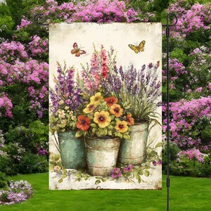 May include: A garden flag with a watercolour painting of three metal buckets filled with colourful flowers. The flowers include yellow, red, and purple blooms. Two butterflies are depicted above the buckets. The flag is displayed in a lush green garden.
