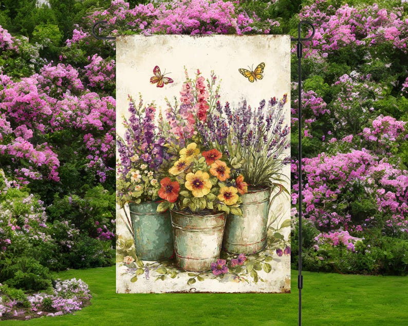 May include: A garden flag with a watercolour painting of three metal buckets filled with colourful flowers. The flowers include yellow, red, and purple blooms. Two butterflies are depicted above the buckets. The flag is displayed in a lush green garden.
