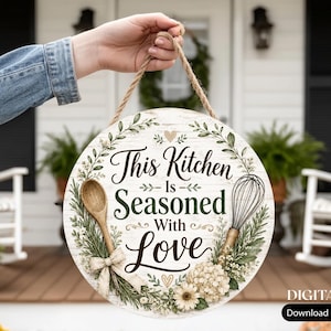 May include: A round wooden sign with a white wood grain background featuring the text This Kitchen Is Seasoned With Love in black script and serif fonts. The design includes illustrations of a wooden spoon, a wire whisk, and floral greenery.