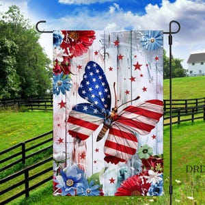 May include: A garden flag featuring a butterfly with wings designed as the American flag, set against a white wooden background with red stars and floral accents. The flag is displayed outdoors with a green lawn and blue sky.