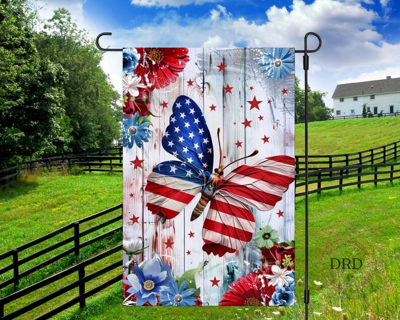 May include: A garden flag featuring a butterfly with wings designed as the American flag, set against a white wooden background with red stars and floral accents. The flag is displayed outdoors with a green lawn and blue sky.