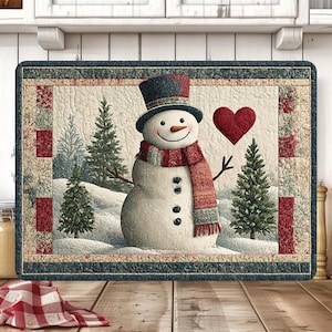 Puede incluir: Un mantel individual rectangular acolchado con un muñeco de nieve con sombrero de copa, bufanda y un corazón rojo. El mantel individual tiene una escena invernal con pinos y suelo cubierto de nieve. El borde es un patrón azul y rojo.