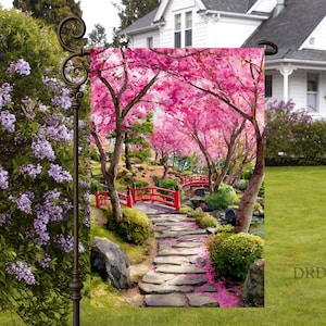 May include: A garden flag featuring a vibrant scene of a Japanese garden. The image showcases a stone pathway leading to a red bridge, surrounded by pink cherry blossom trees. The flag is designed for outdoor use and adds a touch of nature to any yard.