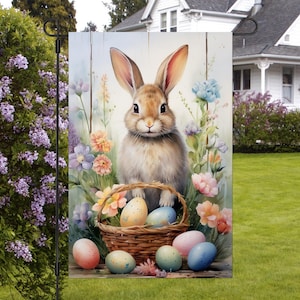 May include: A brown bunny rabbit with long ears sits in a basket filled with colourful Easter eggs. The basket is surrounded by spring flowers, including pink, purple, and yellow blooms. The bunny is looking at the camera with a sweet expression.