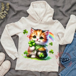 May include: A white hoodie with a cute cartoon cat holding a pot of gold and a shamrock. The cat has orange and white fur and green eyes. A rainbow is behind the cat.