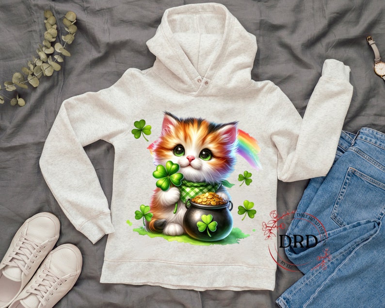 May include: A white hoodie with a cute cartoon cat holding a pot of gold and a shamrock. The cat has orange and white fur and green eyes. A rainbow is behind the cat.