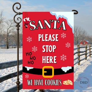May include: A red Christmas garden flag with the words "SANTA PLEASE STOP HERE WE HAVE COOKIES." The flag has a Santa belt design with snowflakes and a Santa illustration. The flag is hanging on a black metal pole in a snowy outdoor scene.