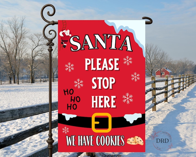 May include: A red Christmas garden flag with the words "SANTA PLEASE STOP HERE WE HAVE COOKIES." The flag has a Santa belt design with snowflakes and a Santa illustration. The flag is hanging on a black metal pole in a snowy outdoor scene.