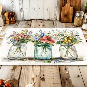 May include: A rectangular kitchen mat featuring a watercolor painting of three glass mason jars filled with colorful wildflowers. The jars are set against a white background, with a rustic wooden table in the foreground.