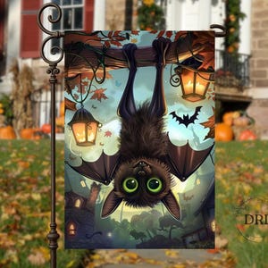 May include: A Halloween garden flag featuring a cartoon bat hanging upside down from a tree branch. The bat has large green eyes and is surrounded by lanterns, bats, and a spooky house scene. The flag is displayed outdoors.