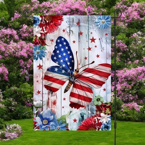 May include: A garden flag featuring a butterfly with wings designed as the American flag. The flag has a white wooden background with red stars and floral accents in red, white, and blue. The flag is displayed in a garden.