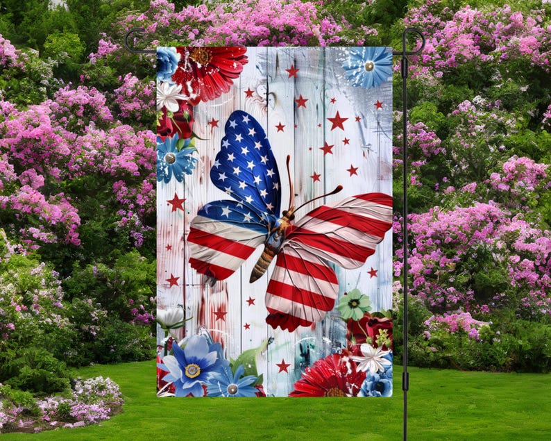 May include: A garden flag featuring a butterfly with wings designed as the American flag. The flag has a white wooden background with red stars and floral accents in red, white, and blue. The flag is displayed in a garden.