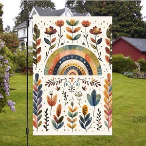 Puede incluir: Bandera de jardín decorativa con un diseño de arcoíris y floral. La bandera tiene un fondo crema y se muestra al aire libre. El diseño incluye un arcoíris, flores y elementos botánicos.