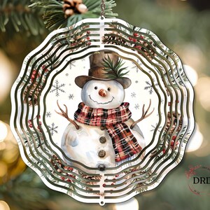Op de afbeelding: Een gelaagde, cirkelvormige kerstversiering met een aquarel sneeuwpop met een hoge hoed en een geruite sjaal. De versiering heeft een decoratieve rand met dennennaalden, bessen en sneeuwvlokken. Het hangt aan een ketting.