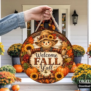 May include: A round wooden sign featuring a scarecrow illustration, sunflowers, pumpkins, and autumn leaves. The text reads Welcome FALL Y'all. The sign has a rustic wood-plank background and is suspended by a thick, light brown jute rope hanger.