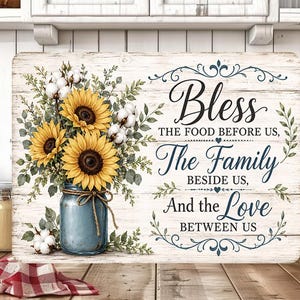 Puede incluir: Un letrero de pared rectangular con fondo de madera blanca que muestra un frasco azul lleno de girasoles amarillos y flores de algodón blancas. El texto dice: Bless THE FOOD BEFORE US, The Family BESIDE US, And the Love BETWEEN US.