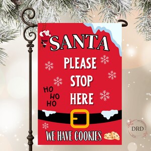 May include: A red Christmas garden flag with the words "SANTA PLEASE STOP HERE" in white. The flag features snowflakes, Santa Claus, and a belt design with the text "WE HAVE COOKIES".