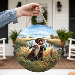 Könnte beinhalten: Runder Holzschild mit einem braunen Labrador Retriever, der eine Ente im Maul hält. Die Szene zeigt eine ruhige Landschaft mit See, Schilf und fliegenden Enten. Ein Juteseil ist zum Aufhängen befestigt. Durchmesser ca. 25 cm.