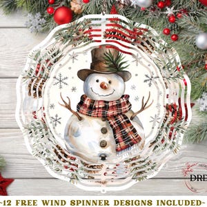 Op de afbeelding: Een witte, cirkelvormige windspinner met een aquarel sneeuwpop met een hoge hoed en een geruite sjaal. Het ontwerp bevat sneeuwvlokken en is omgeven door een decoratieve rand met dennentakken en rode accenten. De tekst onderaan luidt "~12 FREE WIND SPINNER DESIGNS INCLUDED~".