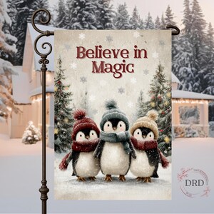May include: A garden flag features three penguins wearing winter hats and scarves, standing in a snowy scene. The flag reads "Believe in Magic" in red text, with a wintery background of trees and a house.