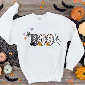May include: White sweatshirt with a graphic of the word "BOO" in colorful, patterned letters. A skeleton is holding a letter "O" and a black cat is sitting below the word.