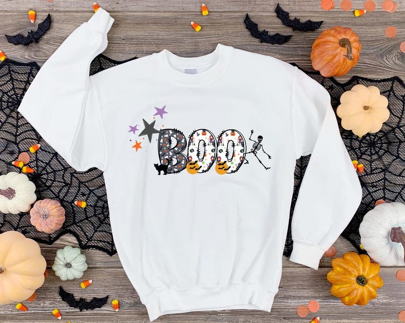 May include: White sweatshirt with a graphic of the word "BOO" in colorful, patterned letters. A skeleton is holding a letter "O" and a black cat is sitting below the word.