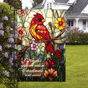 May include: A stained glass style garden flag with a red cardinal bird perched on a branch. The flag has a green, blue, and yellow background with red, yellow, and white flowers. The text on the flag reads "Angels appear when Cardinals are near".