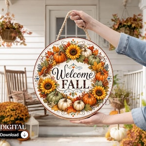 May include: A round wooden sign featuring a decorative wreath of sunflowers, orange and green pumpkins, and autumn leaves. The centre of the sign displays the text "Welcome FALL" in a black script and serif font, suspended by a thick rope.