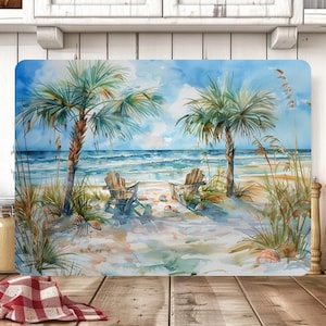 May include: Rectangular cutting board featuring a watercolor beach scene. The image depicts two palm trees, two wooden chairs, and the ocean under a blue sky. The board is likely made of glass or a similar material.