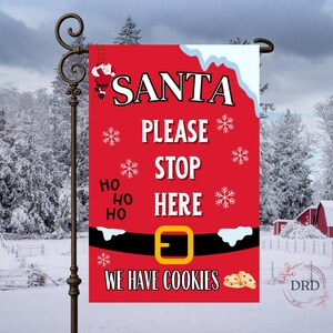 May include: A red Christmas garden flag with the words "SANTA PLEASE STOP HERE" in white. The flag features a Santa belt design with snowflakes and the phrase "WE HAVE COOKIES". A decorative flag for the holiday season.