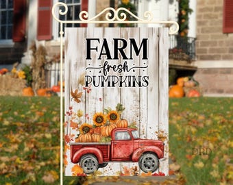 Può includere: Una bandiera da giardino rettangolare con uno sfondo a doghe di legno bianco e il testo nero FARM fresh PUMPKINS. Un furgoncino rosso vintage è carico di zucche arancioni e girasoli gialli, circondato da foglie autunnali sparse.
