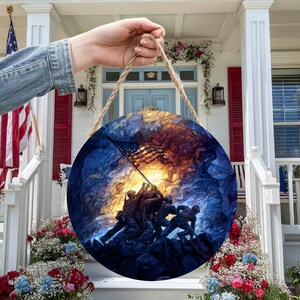 May include: Round wooden wall hanging featuring a patriotic scene of soldiers raising the American flag, with a rope for hanging. The artwork is set against a backdrop of blue and gold hues, evoking a sense of heroism and national pride.