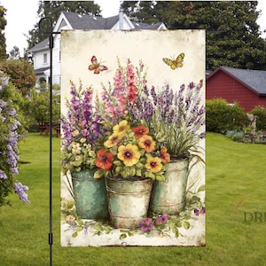 May include: A garden flag featuring a floral still life with three metal pots overflowing with colorful flowers. Butterflies flutter above the blooms. The flag is displayed outdoors in a yard.
