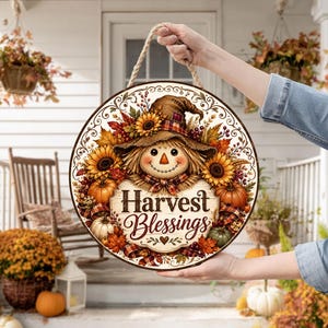 May include: A round wooden sign featuring a scarecrow illustration, sunflowers, pumpkins, and autumn leaves. The text Harvest Blessings is printed in the centre. The sign includes a thick rope hanger attached to the top for display purposes.
