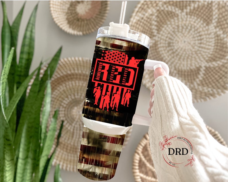 May include: A stainless steel tumbler with a black and red design. The tumbler features a red, white, and blue American flag with the words "Remember Everyone Deployed" and "RED" in red text. The tumbler has a white lid and a white handle.