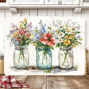 May include: A rectangular cutting board featuring a watercolor painting of three mason jars filled with colorful wildflowers. The jars are clear, and the flowers include red, yellow, purple, and white blooms. The background is a soft, neutral tone.