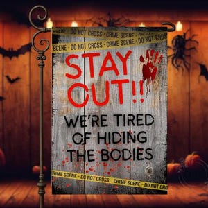 May include: A Halloween garden flag with a weathered wood background. The flag reads "STAY OUT!!! WE'RE TIRED OF HIDING THE BODIES" in red and black text. A red handprint and yellow crime scene tape complete the design.