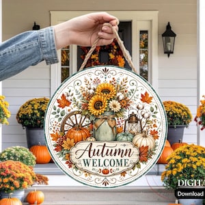 May include: A round wooden sign featuring a printed illustration of sunflowers in a mason jar, pumpkins, a lantern, and a wagon wheel. The text Autumn WELCOME is centred in a decorative frame. The sign hangs from a thick jute rope.