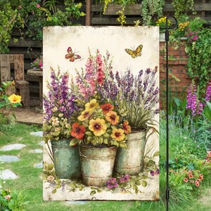 May include: A garden flag with a watercolour painting of colourful flowers in metal pots. The flowers include purple, pink, yellow, and orange blooms. Two butterflies are depicted above the flowers. The flag is displayed in a garden setting.